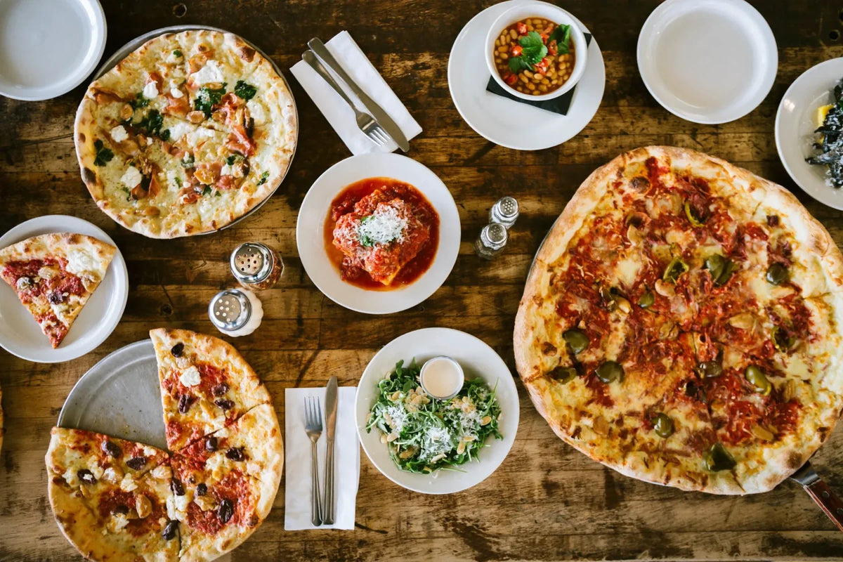 Pizzas and plates of food spread across a wooden table.